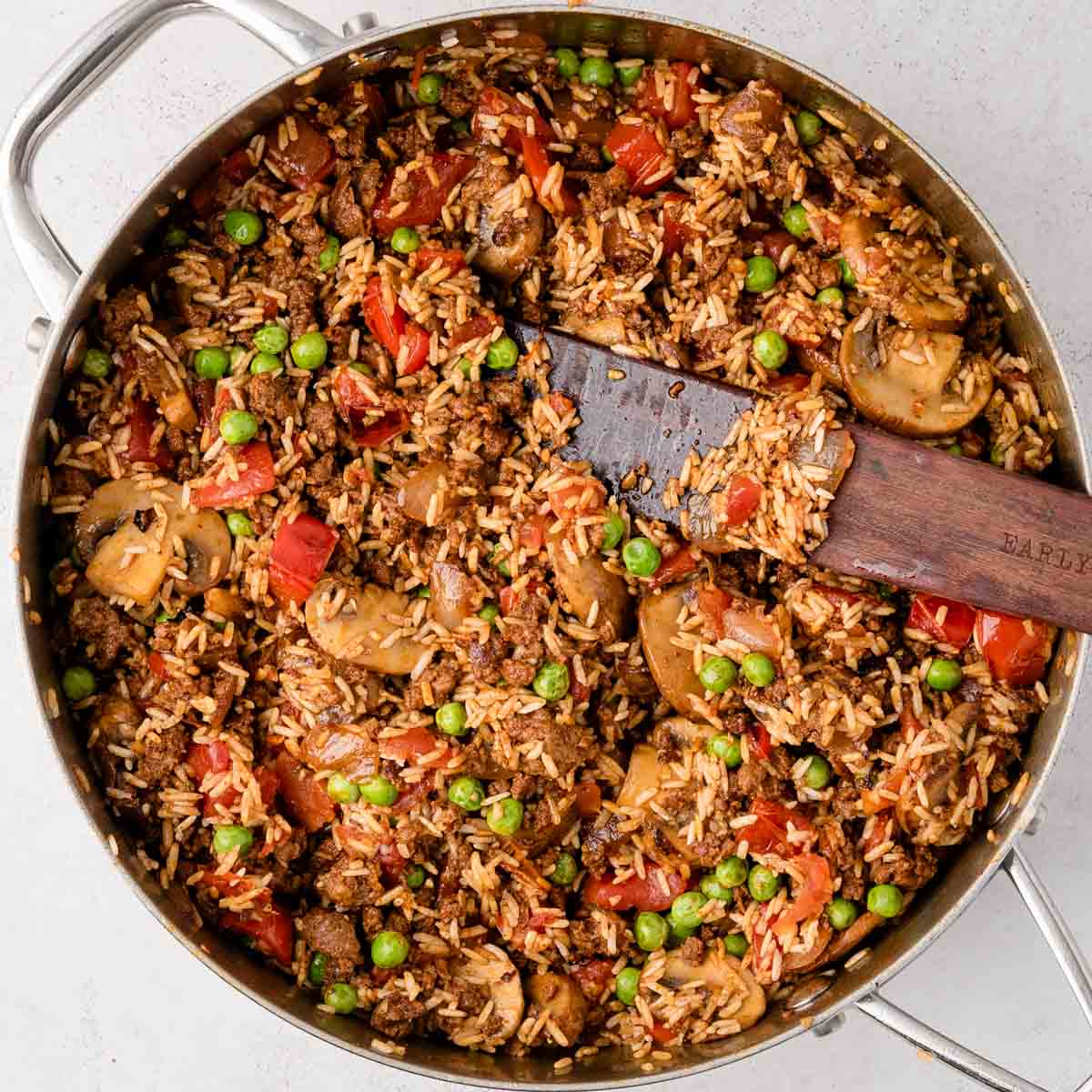 A large skillet filled with rice, ground meat, mushrooms, peas, tomatoes, and a wooden spatula.