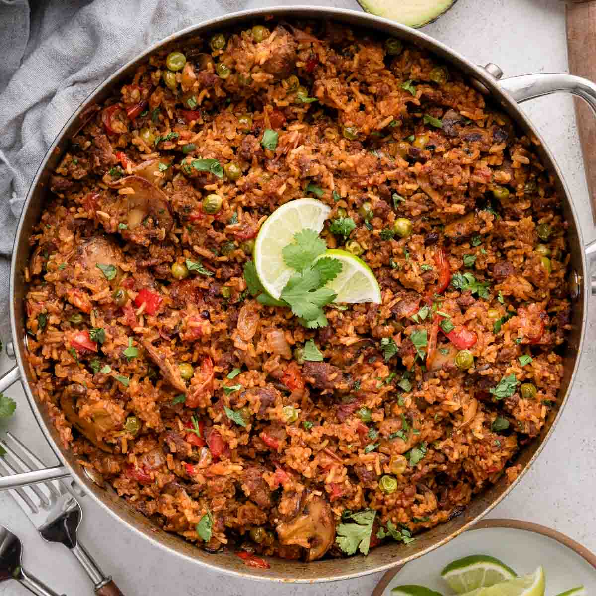A skillet recipe of seasoned chorizo rice with vegetables and peas, garnished with lime wedges and cilantro.