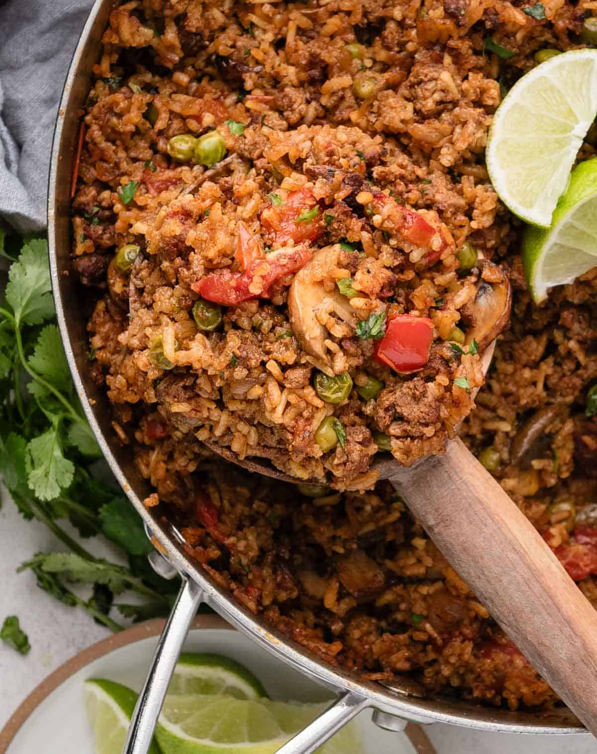 A wooden spoon scoops seasoned chorizo rice with ground meat, peas, and tomatoes from a pan, garnished with lime wedges.
