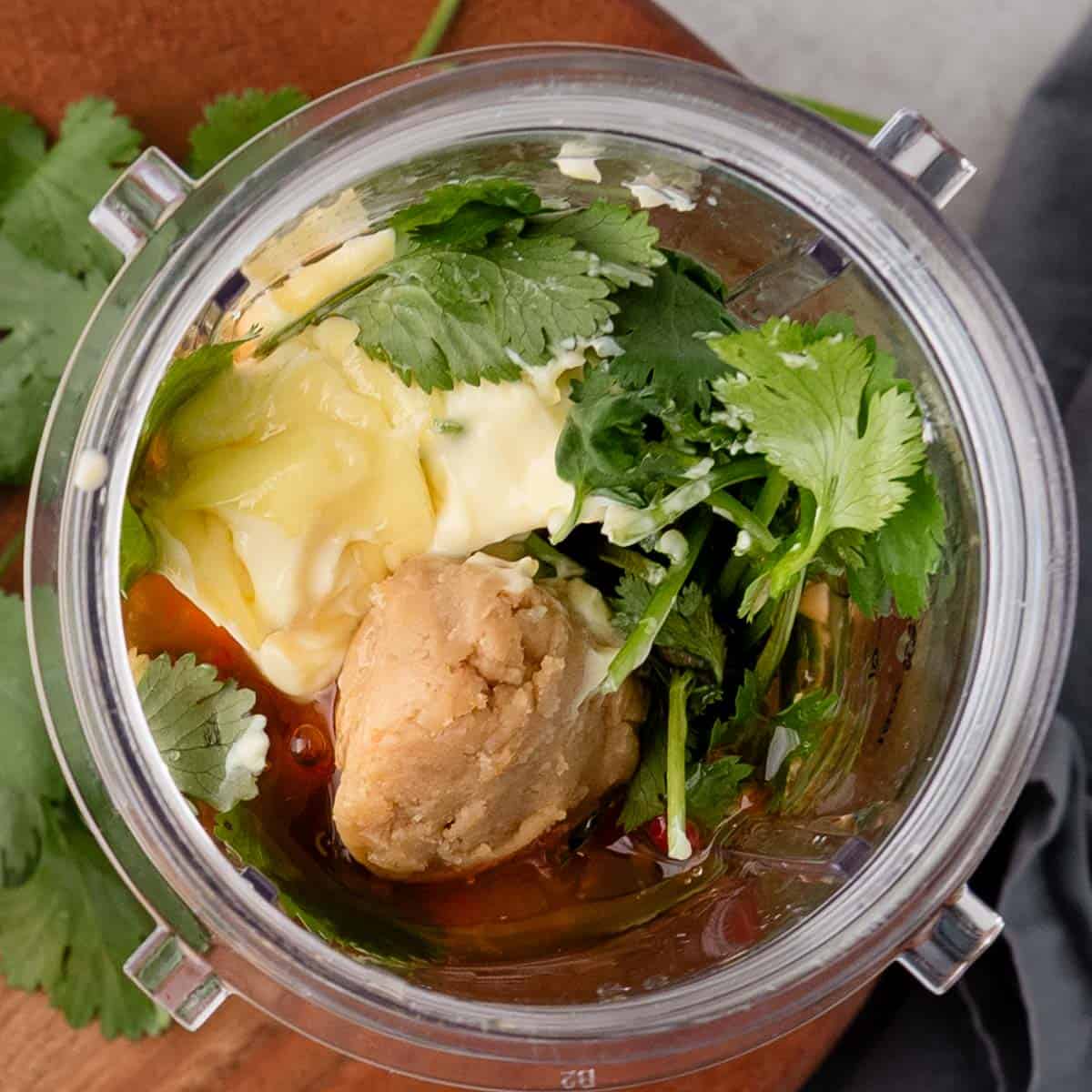 A blender cup filled with cilantro, mayonnaise, miso paste, and sauce, ready to be blended.