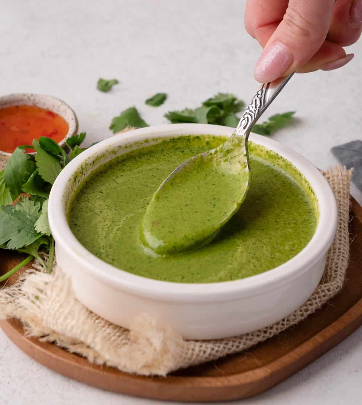 A hand holding a spoon above a bowl of green sauce, with cilantro and another dip in the background.