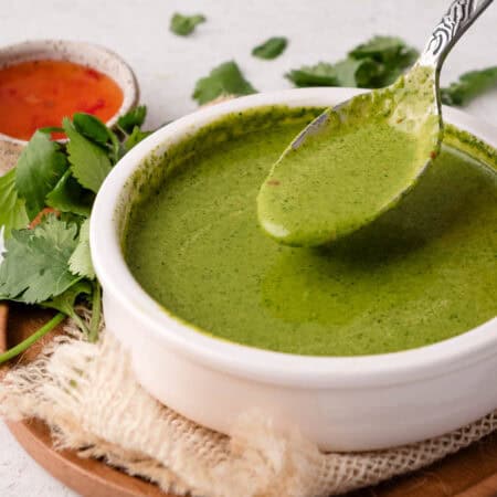 A spoon lifts green cilantro chutney from a white bowl, with herbs and a red sauce nearby.