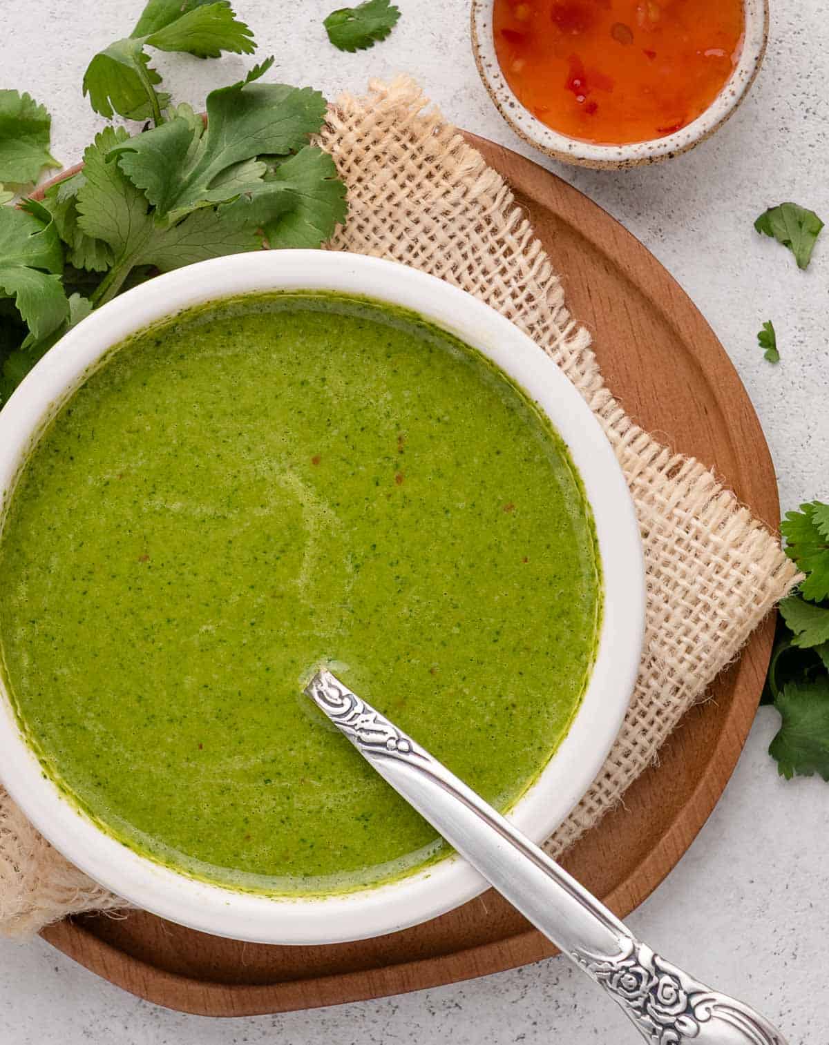 A bowl of green cilantro sauce with a spoon, on a wooden board with fresh cilantro and a small bowl of chili sauce.