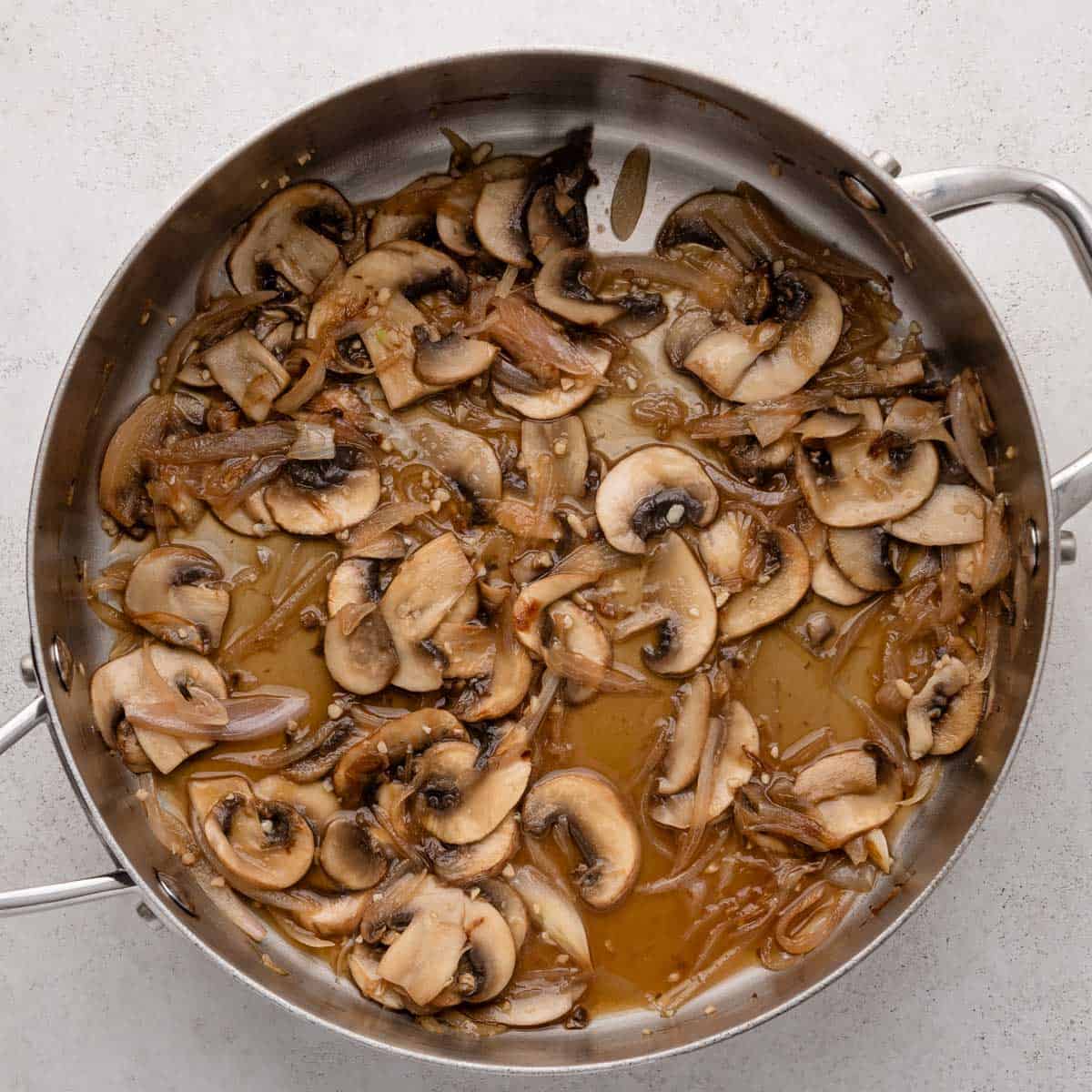 Sliced mushrooms and onions sautéing in a stainless steel pan with brown sauce.