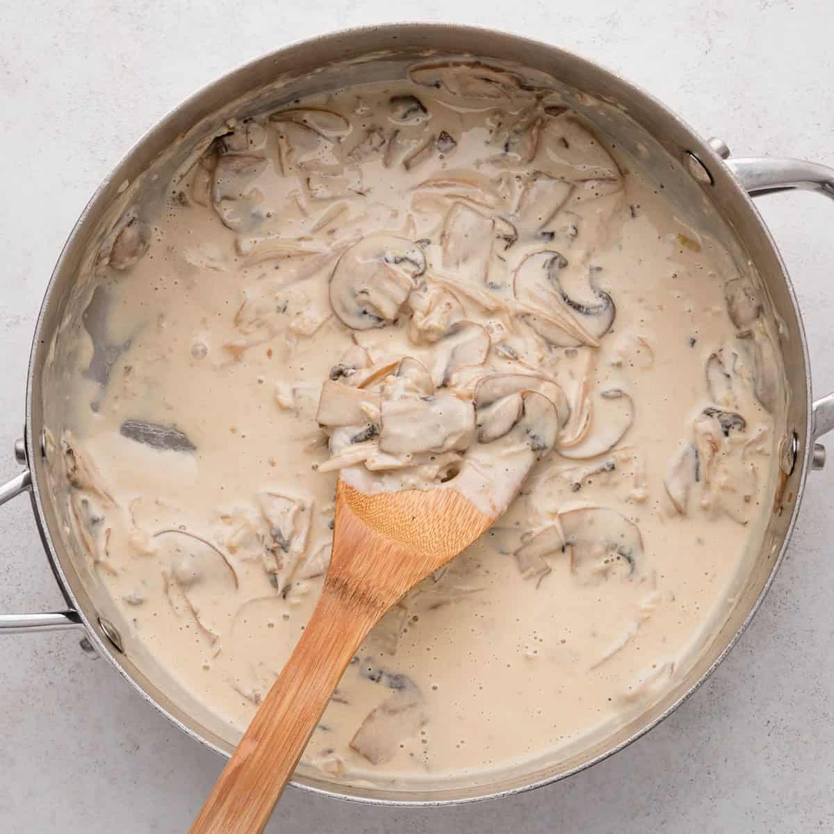 Creamy mushroom sauce with sliced mushrooms in a metal pan, stirred with a wooden spoon.