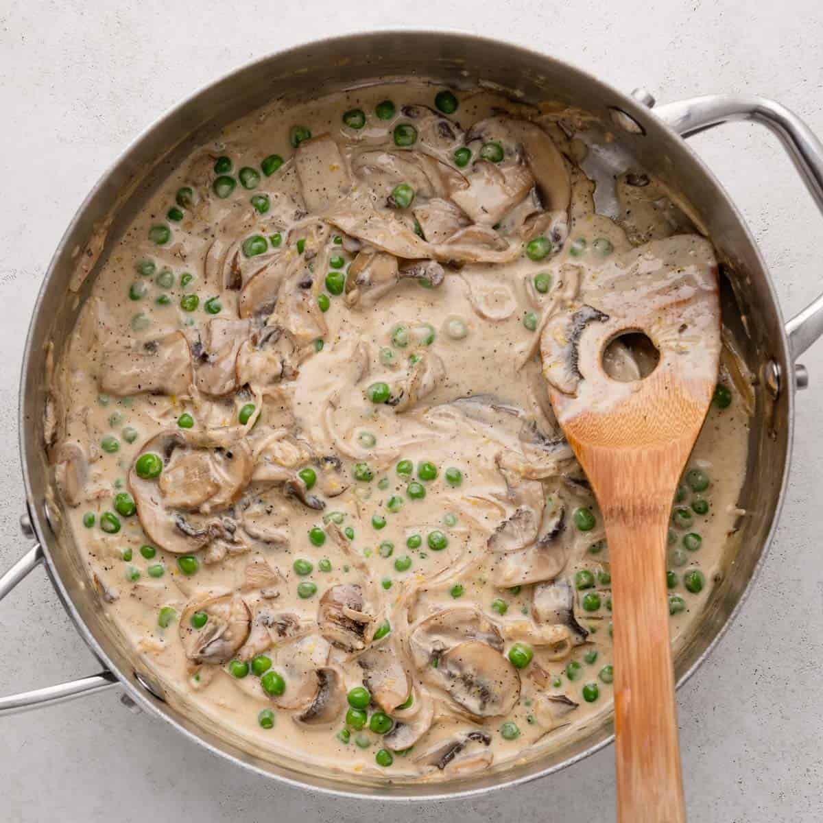 A creamy mushroom and pea sauce in a skillet with a wooden spoon on a light background.