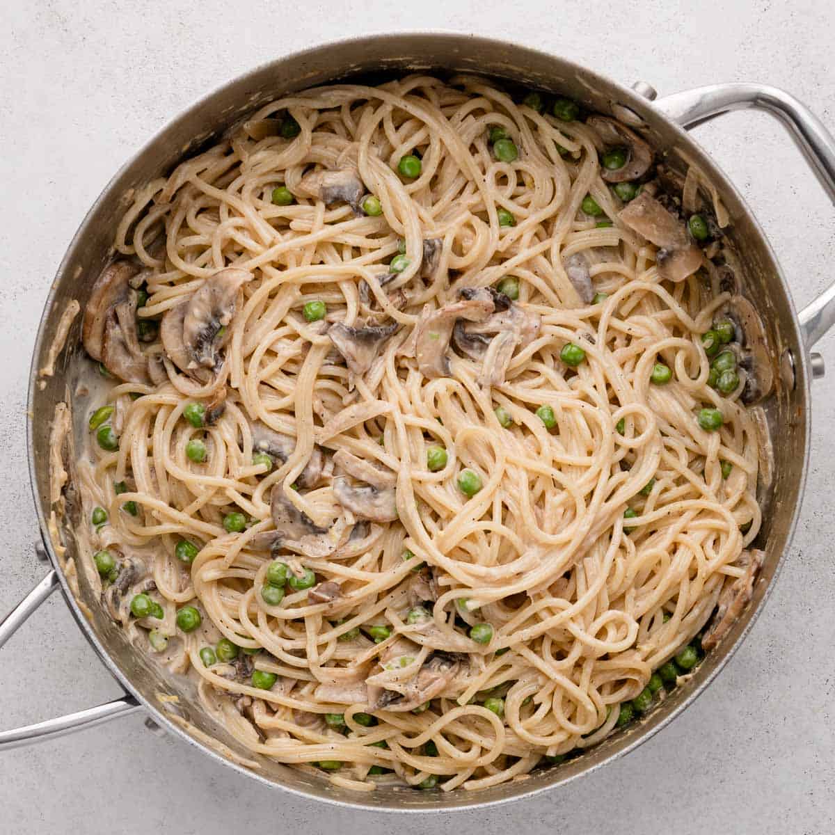 Spaghetti with creamy sauce, mushrooms, and green peas in a metal pan on a light surface.