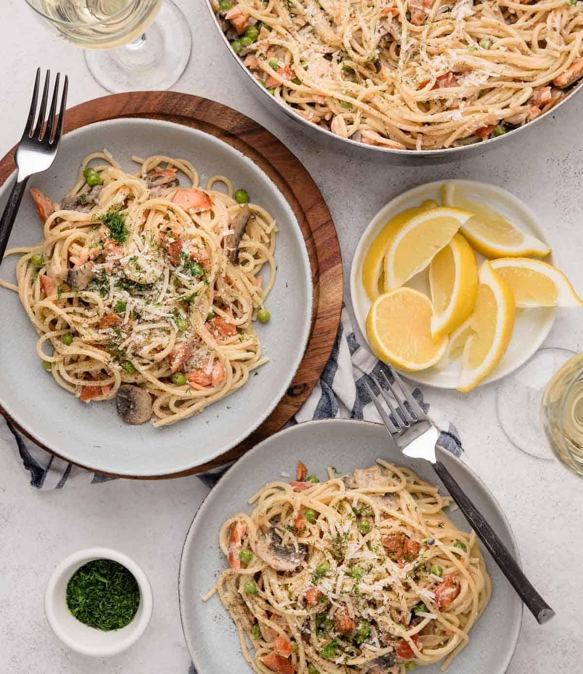 Two plates of creamy pasta with vegetables and parmesan, lemon wedges, and a pot of pasta on a table.