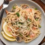Plate of spaghetti with peas, mushrooms, salmon, grated cheese, fresh herbs, and two lemon wedges.