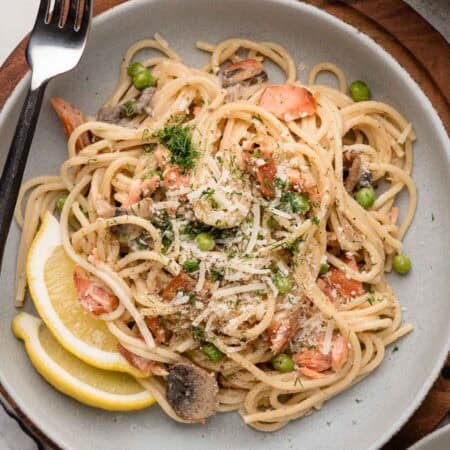Plate of spaghetti with peas, mushrooms, salmon, grated cheese, fresh herbs, and two lemon wedges.