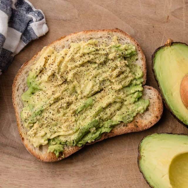 A slice of bread with mashed avocado and black pepper, next to a halved avocado on a wooden surface.
