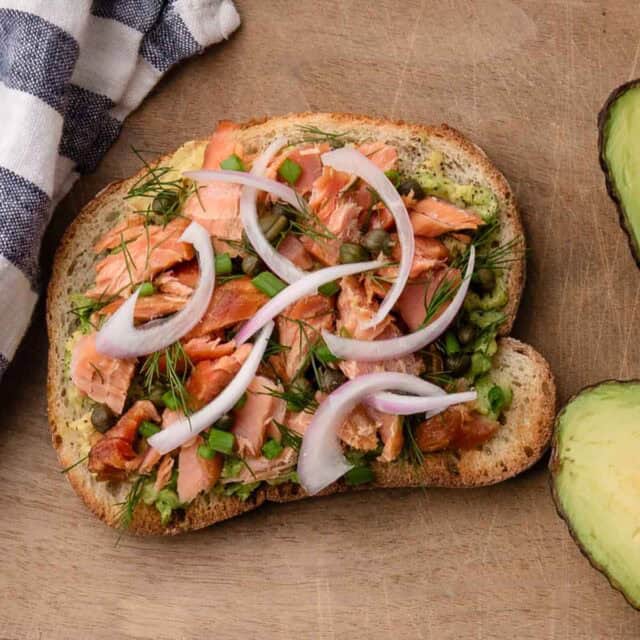 An open-faced sandwich with smoked salmon, red onion, capers, dill, and avocado on rustic bread.