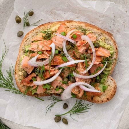 Slice of bread topped with avocado, smoked salmon, sliced onions, capers, and fresh dill on parchment paper.