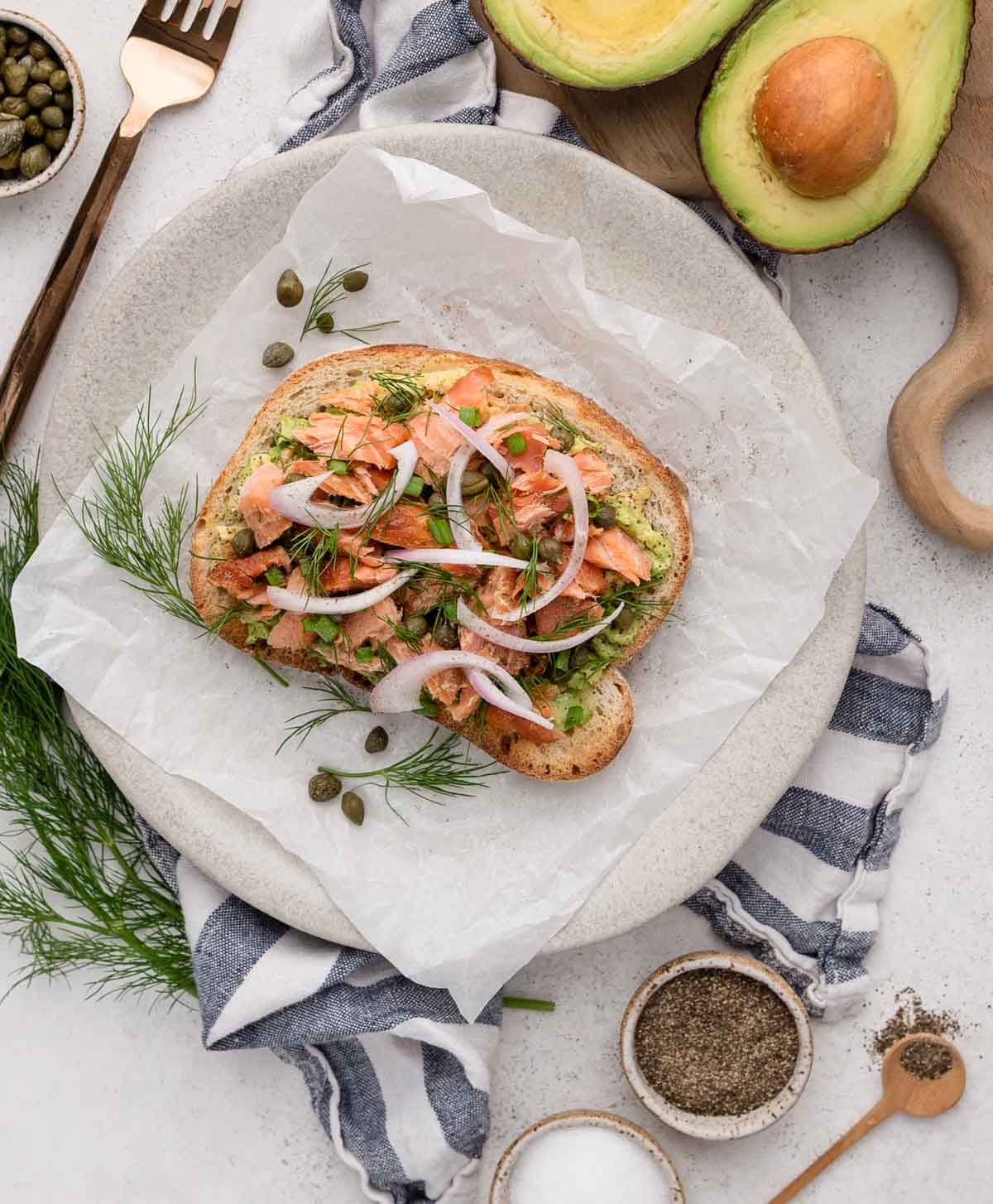 A slice of toast topped with salmon, avocado, onion, dill, and capers on parchment paper.