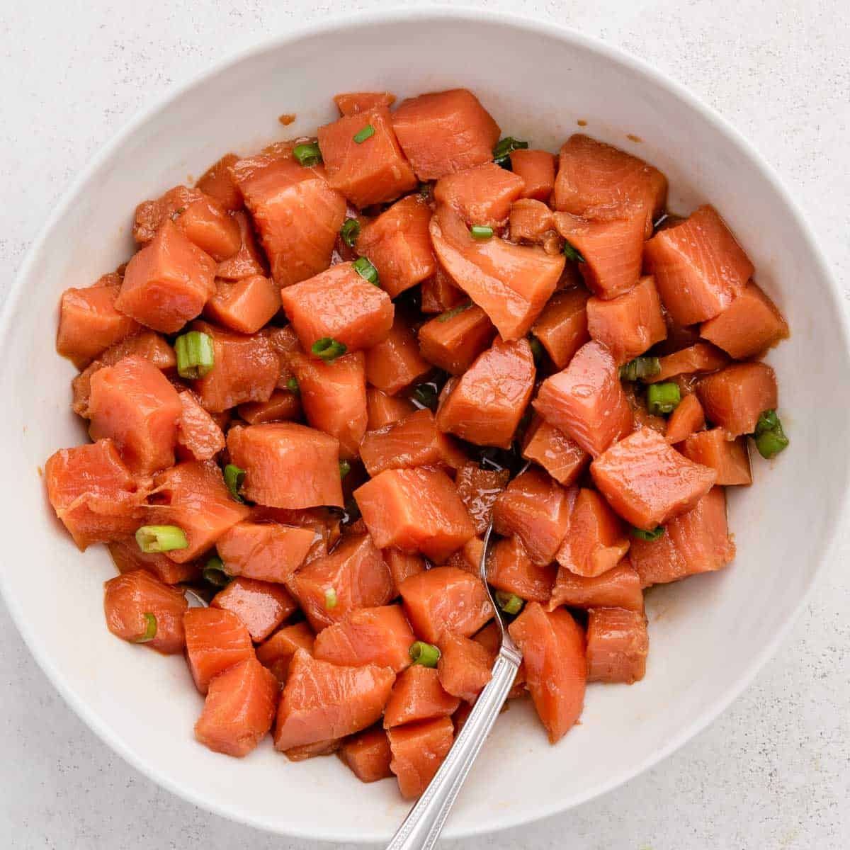A white bowl filled with cubed marinated salmon and chopped green onions, with a spoon inside.