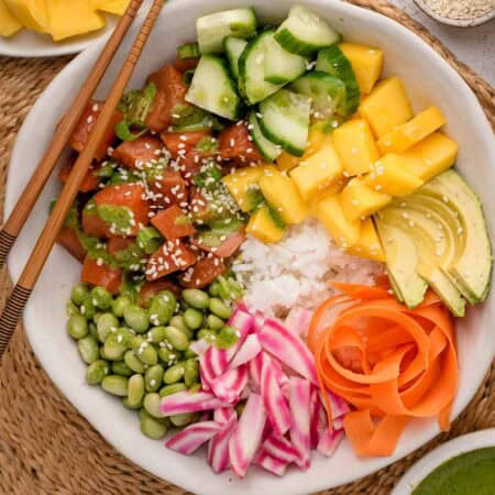 A poke bowl with salmon, cucumber, mango, avocado, edamame, rice, carrot, and radish, topped with sesame seeds.