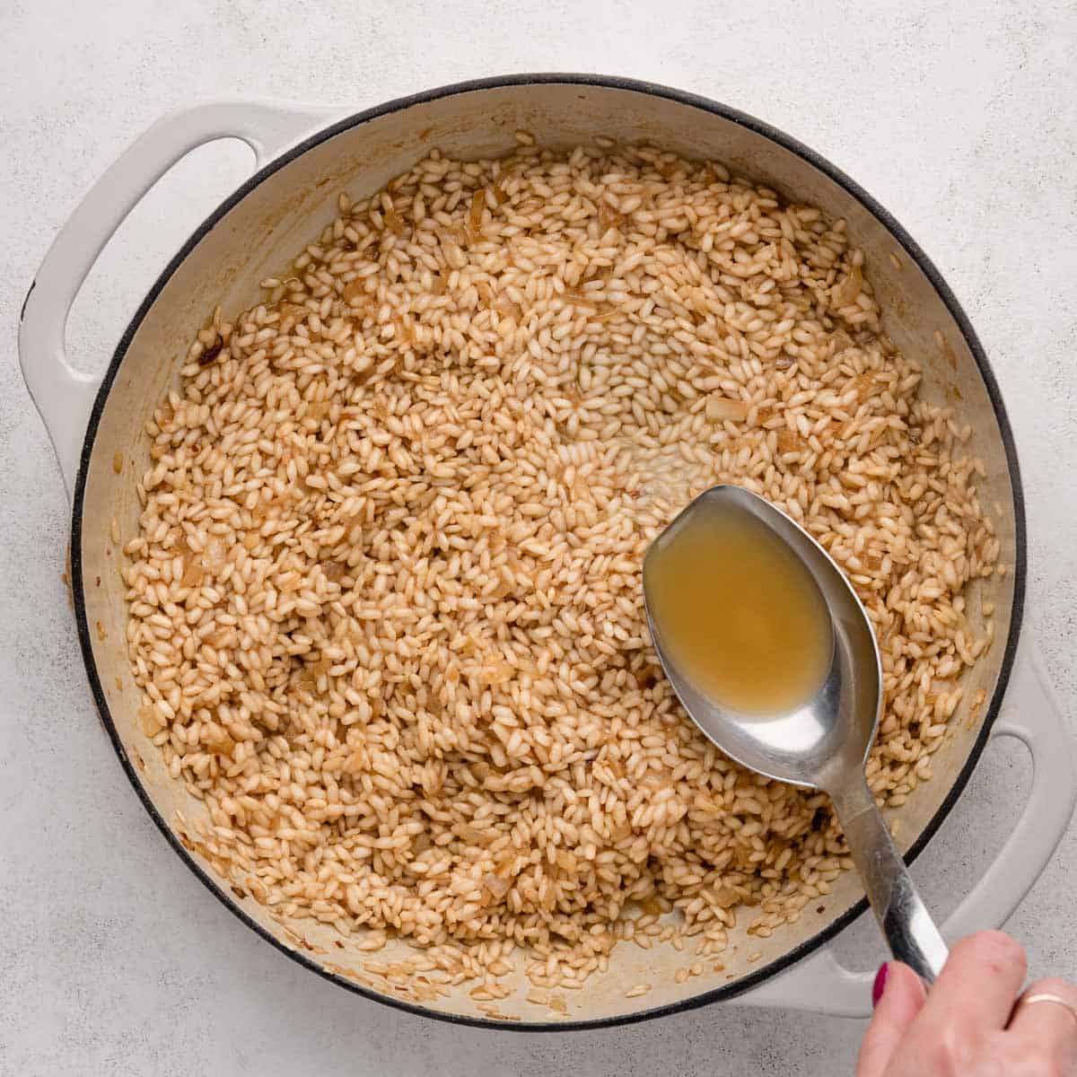 A hand pours broth into a pot of aborio rice on a white surface.