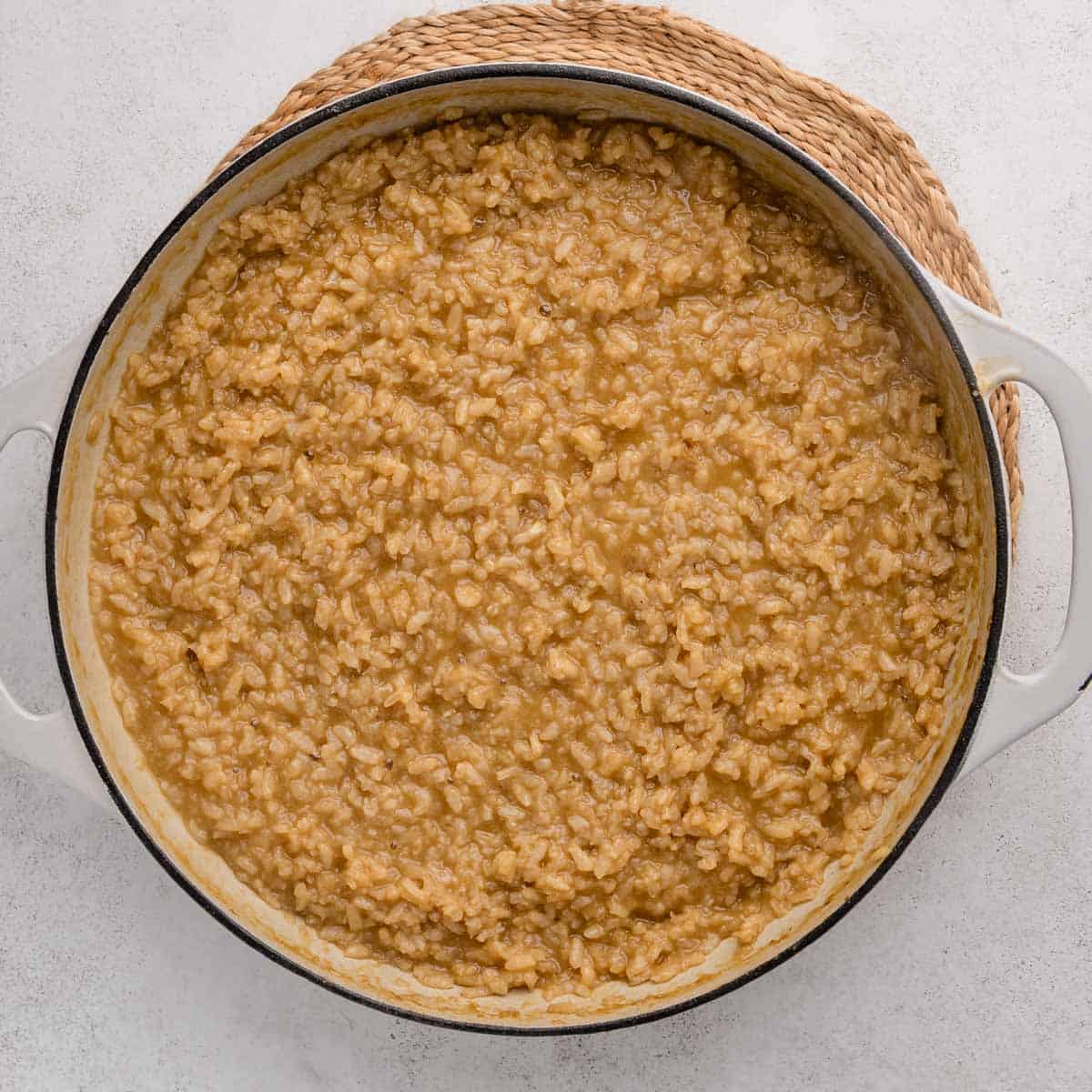 Overhead view of creamy brown risotto in a white pot on a woven mat, on a light surface.