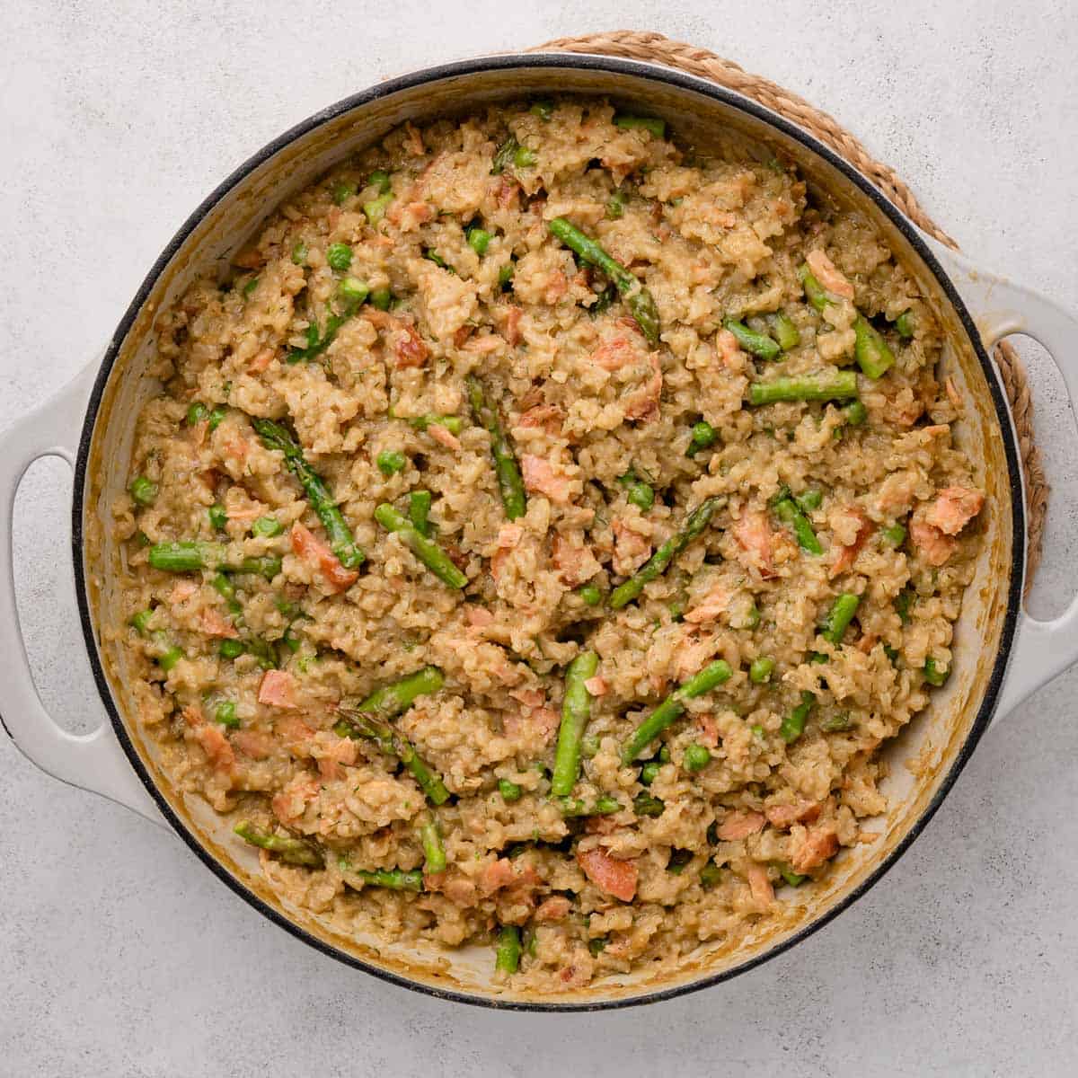 A pot of creamy risotto with asparagus, peas, and chunks of salmon on a light background.