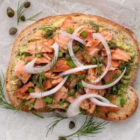 A slice of avocado, smoked salmon toast topped with sliced shallot, capers, chives, and fresh dill.