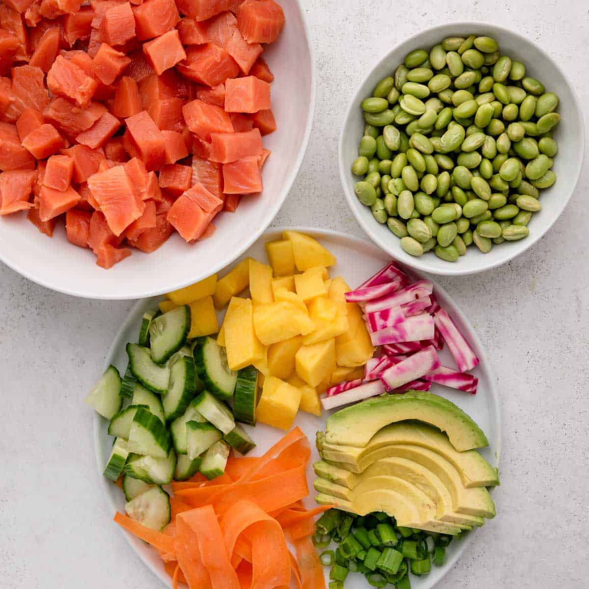 Bowls of cubed raw salmon, edamame, and a plate with cucumber, mango, radish, avocado, carrot, and green onion.