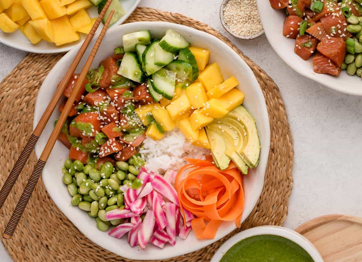 A poke bowl with rice, salmon, mango, cucumber, avocado, edamame, carrots, radish, and sesame seeds.