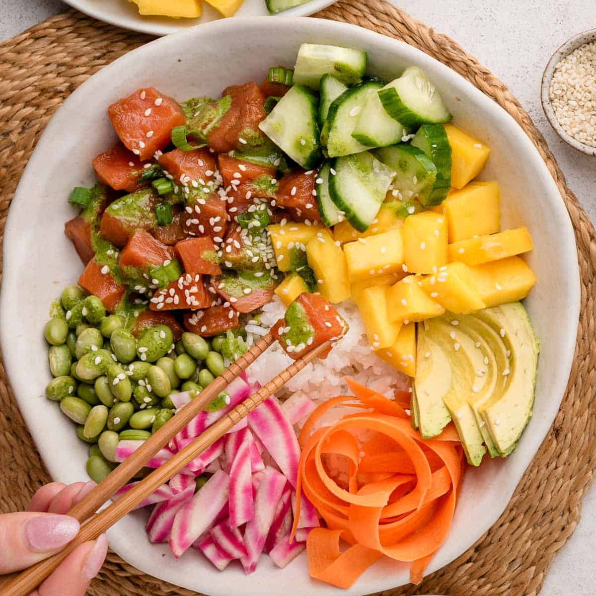 A poke bowl with tuna, cucumber, mango, avocado, edamame, radish, and carrot, sprinkled with sesame seeds.