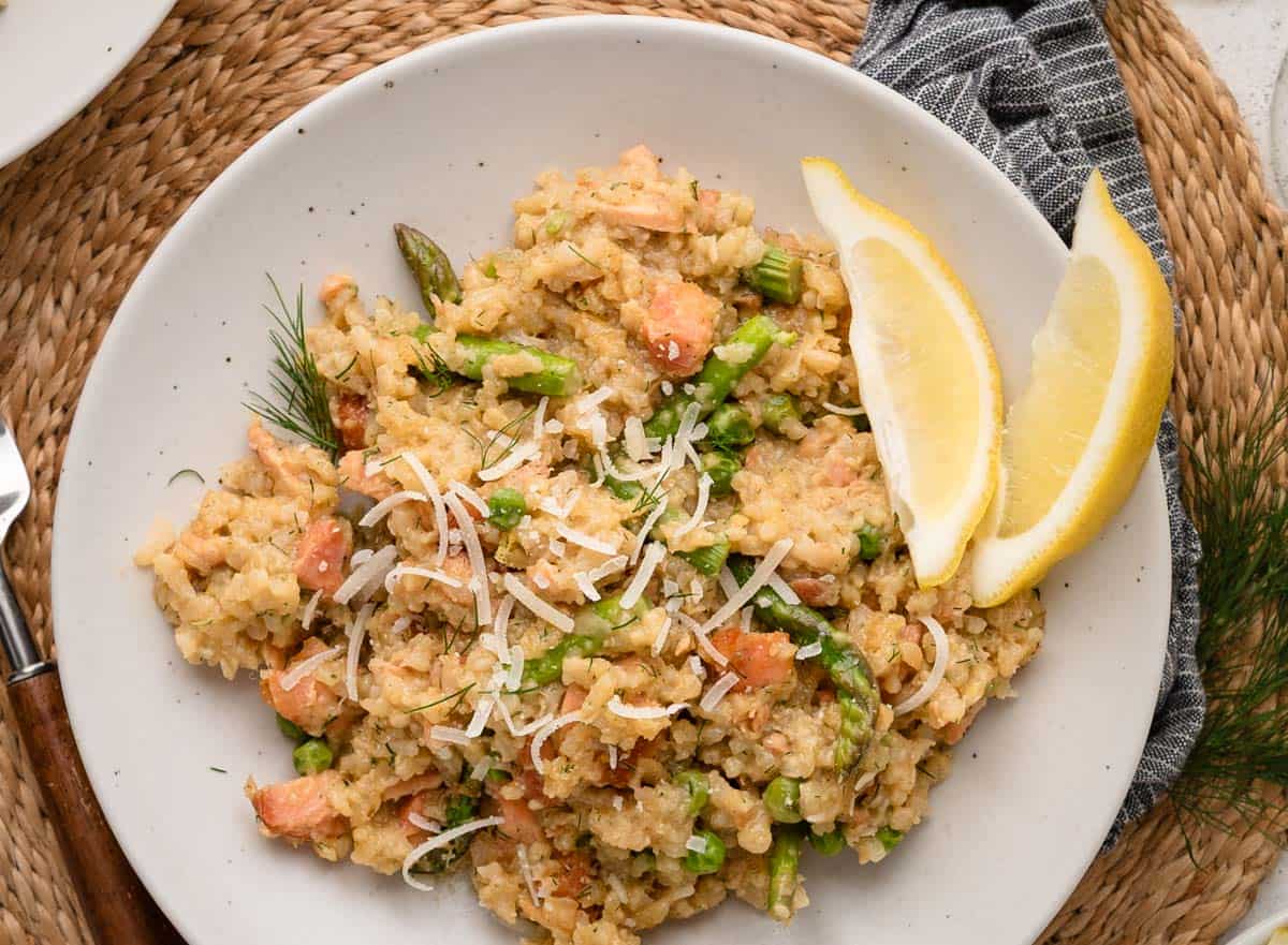 A plate of smoked salmon risotto with vegetables, shredded cheese, and two lemon wedges on the side.
