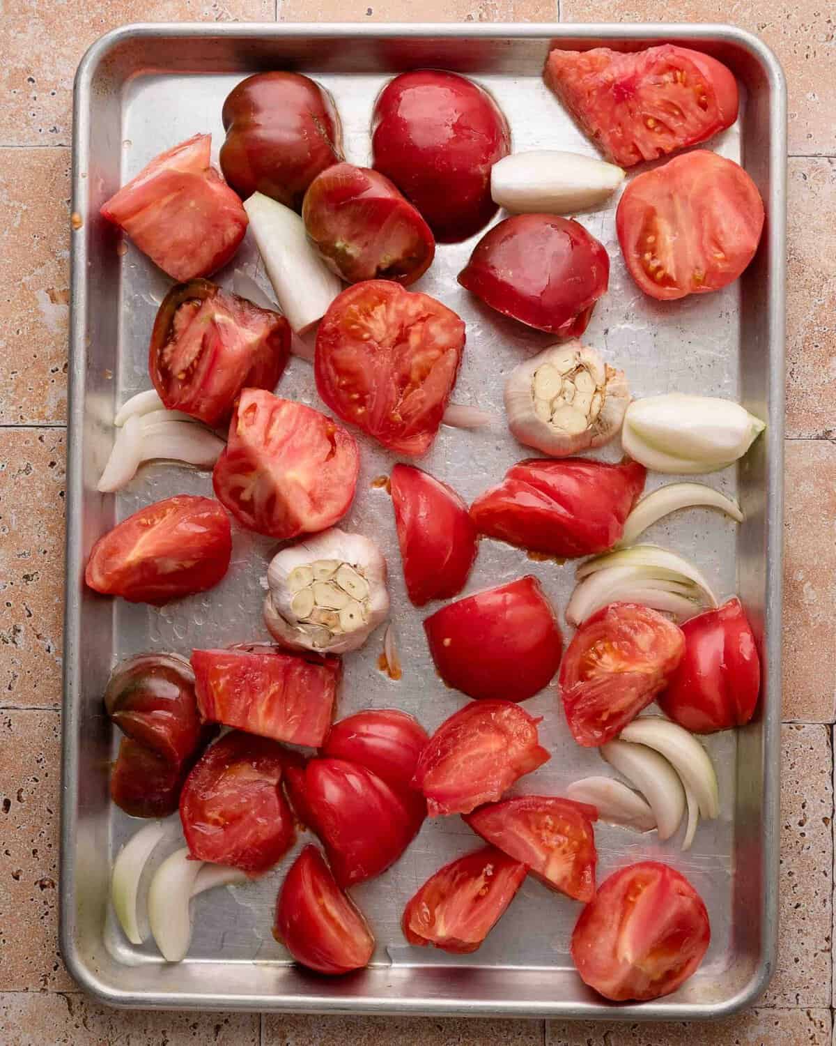 Tomato halves, onion wedges, and garlic bulbs on a metal baking sheet, ready for roasting.