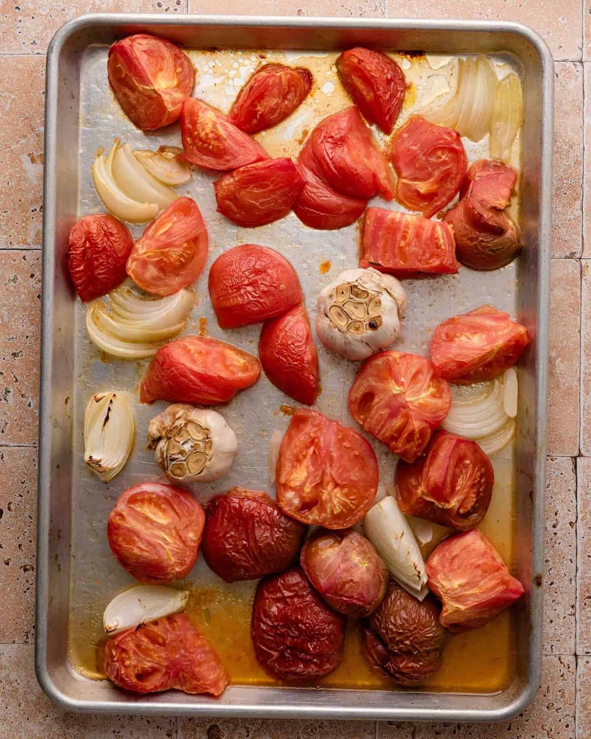 Roasted tomato halves, onion wedges, and halved garlic bulbs on a metal baking sheet.