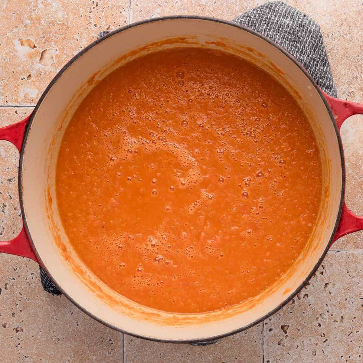 Orange tomato soup simmering in a red pot on a tan tiled countertop with a dark cloth underneath.