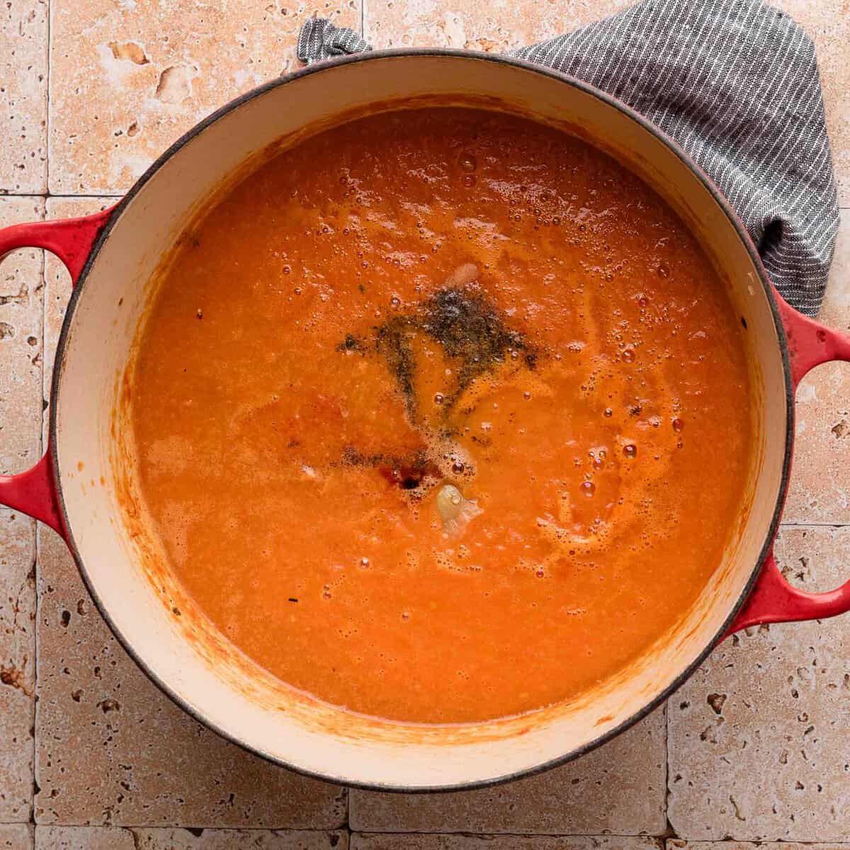 Red tomato soup simmering in a large pot on a tiled countertop, with a towel on the pot handle.