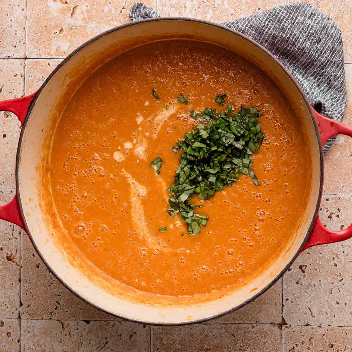 Red pot of tomato soup topped with chopped herbs, placed on a tiled surface with a gray cloth nearby.