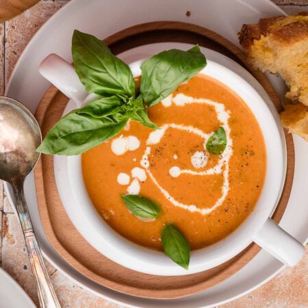 Tomato soup with cream swirl and basil leaves, served with bread on the side and a spoon on the plate.