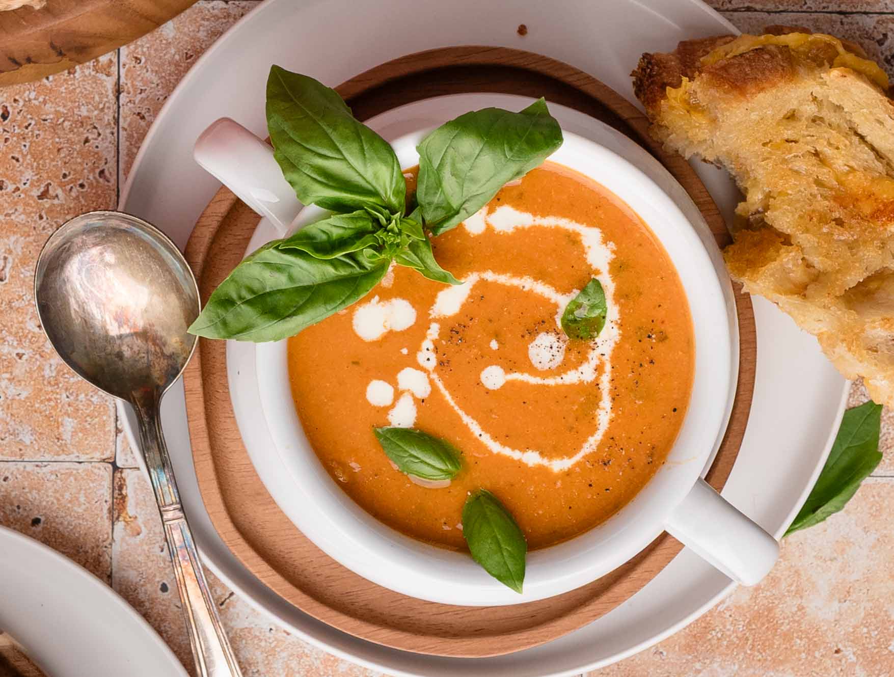 Roasted garlic tomato soup with swirl of cream and basil leaves on top, served with a grilled cheese on the side.