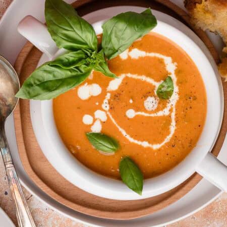 Bowl of roasted garlic tomato soup with cream swirl and basil leaves on top, served with a grilled cheese on the side.