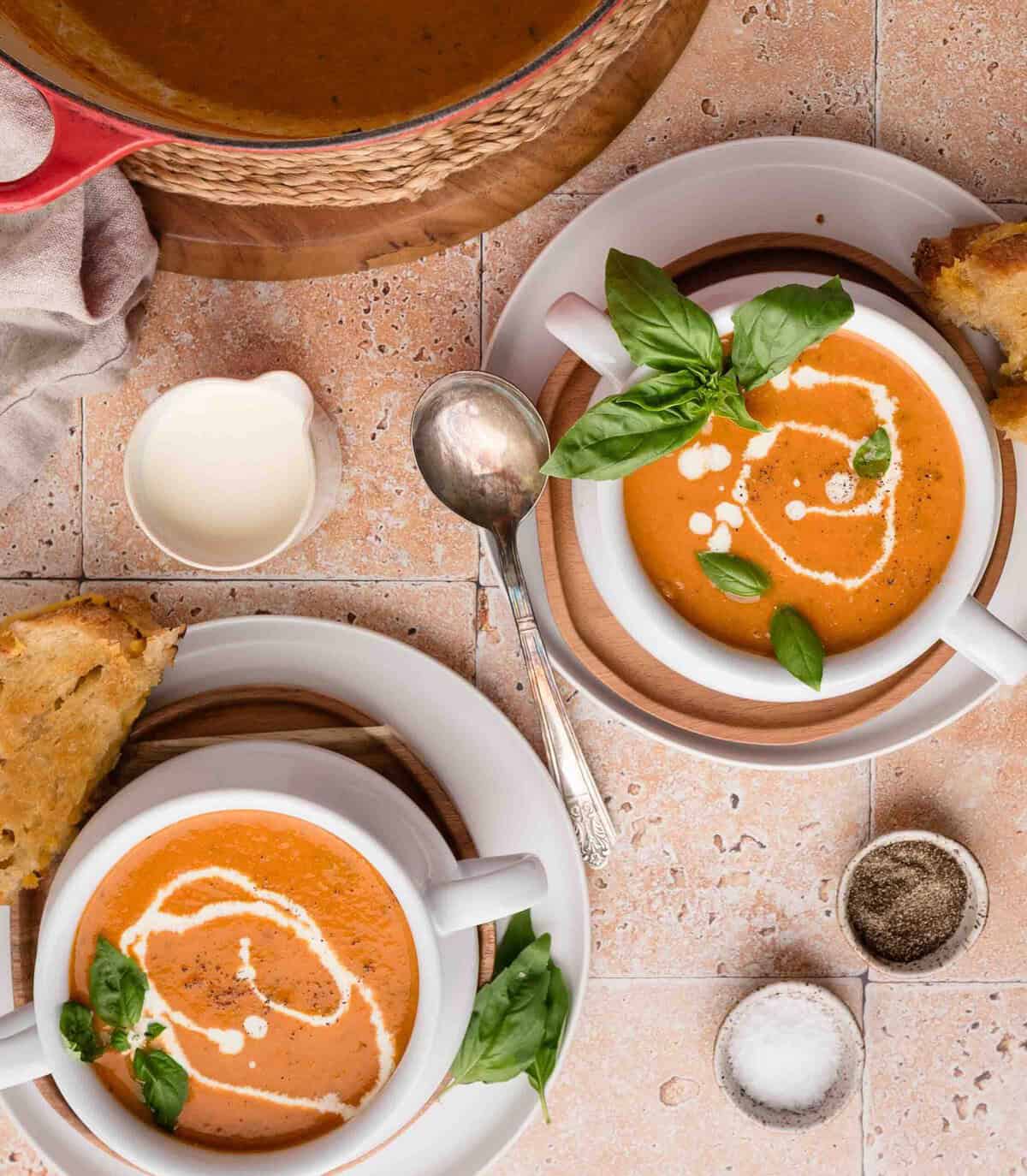 Two bowls of tomato soup with cream and basil garnish, served with bread on a tiled table.