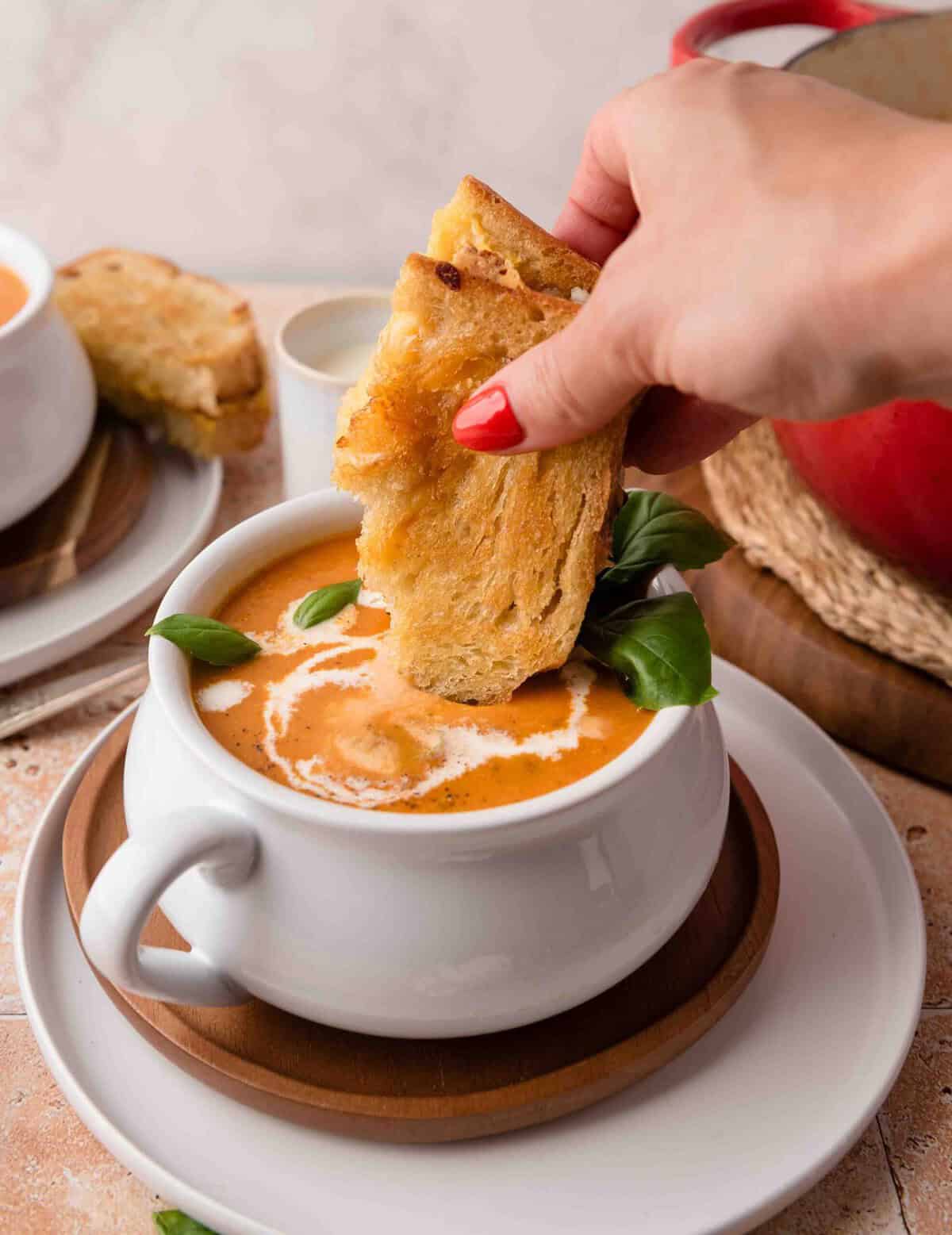 A hand dips toasted bread into creamy roasted garlic tomato soup in a white cup, garnished with herbs.